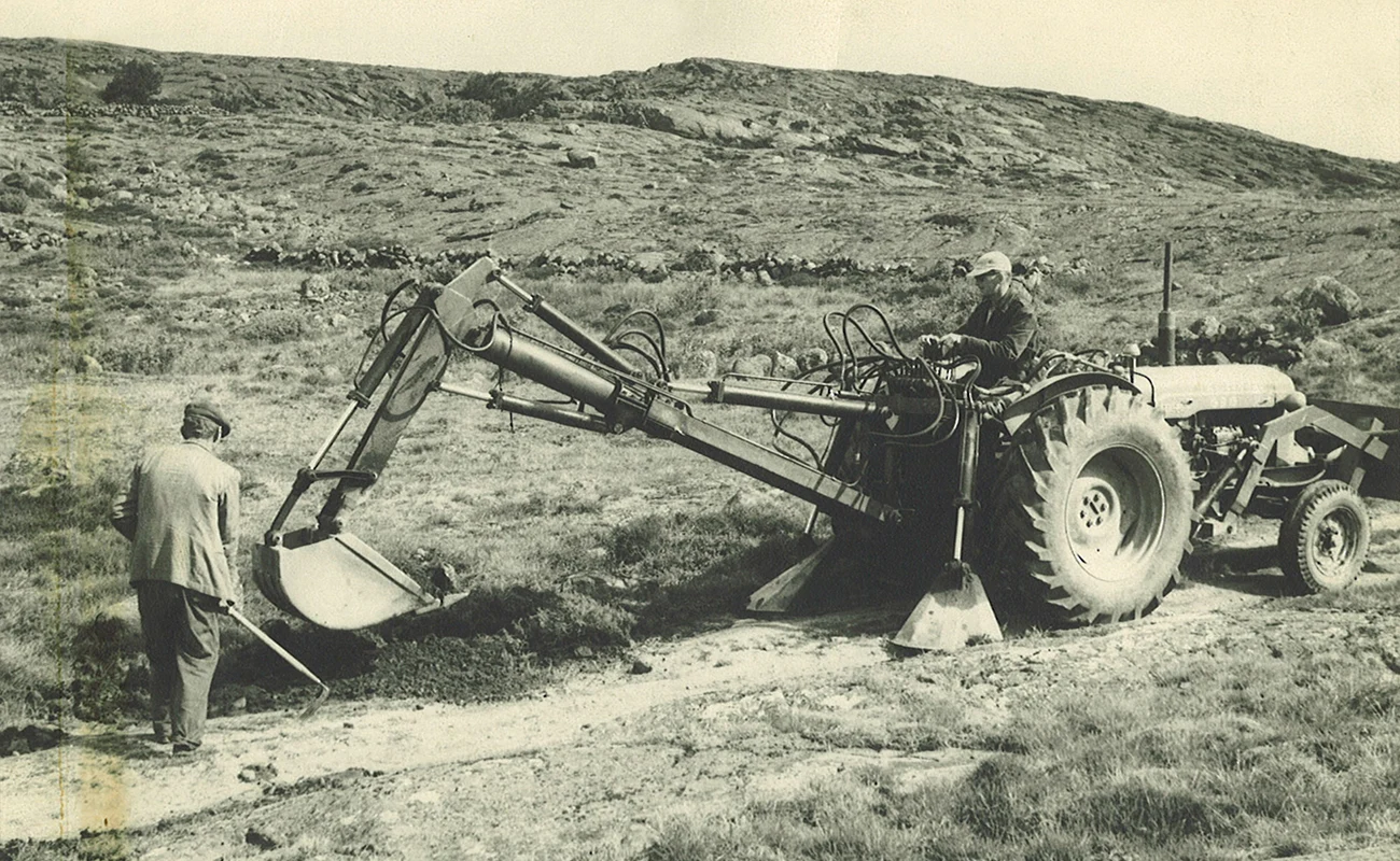 Historiskt svartvitt foto av en traktor med gr&auml;vaggregat som gr&auml;ver dike i bergig terr&auml;ng, med en man som skottar bredvid.