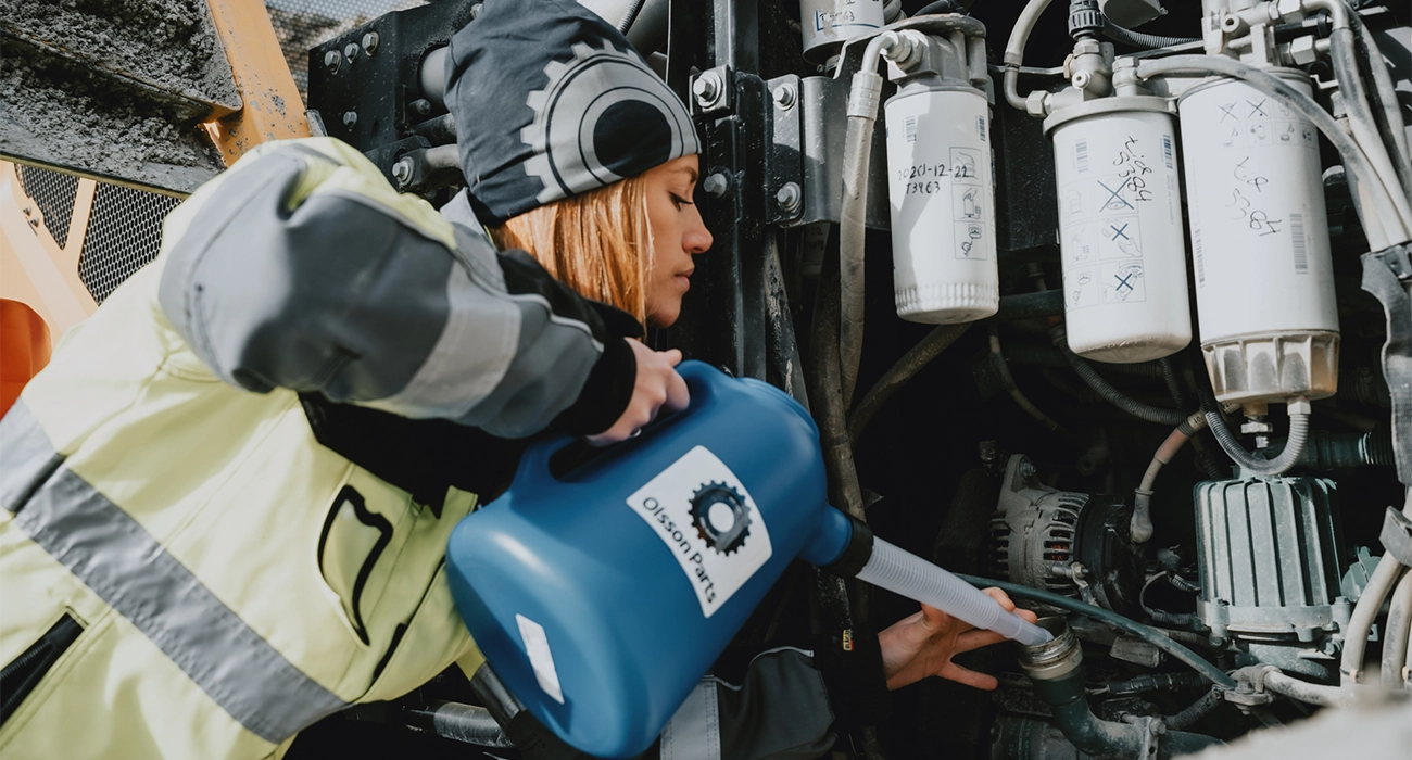 En person i sikkerhedst&oslash;j fylder olie p&aring; en Volvo gummiged med et plastm&aring;leb&aelig;ger fra Olsson Parts.