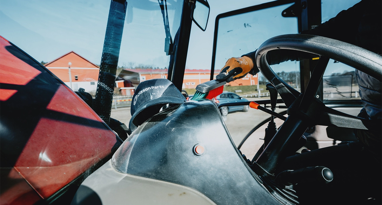 En person børster innsiden av en traktorkabin; en Olsson Parts-caps ligger på dashbordet.