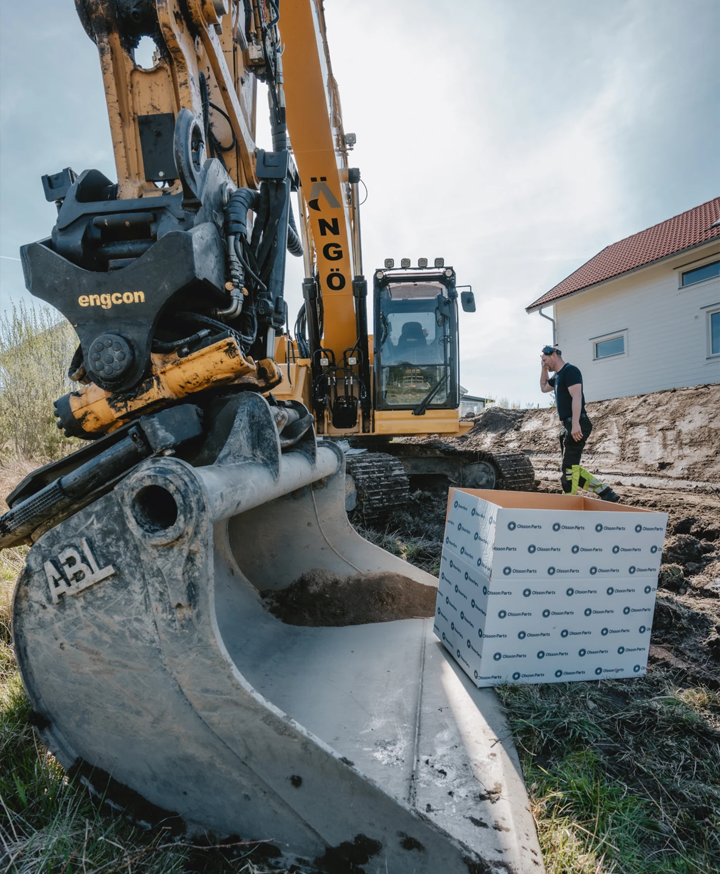 En Caterpillar gravemaskine i arbejde foran et hus med en person ved siden af.