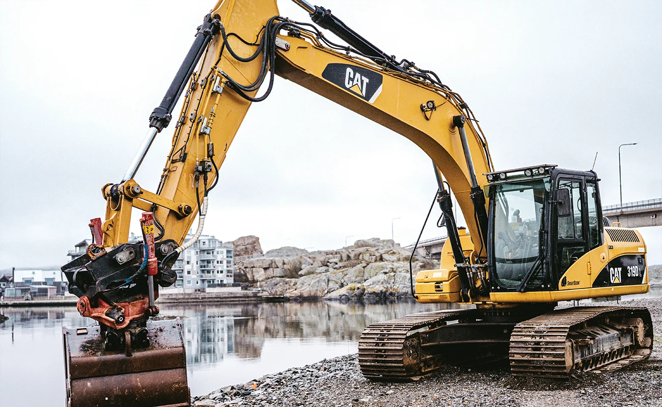 En Caterpillar gravemaskine st&aring;r ved havet med en bro i baggrunden.