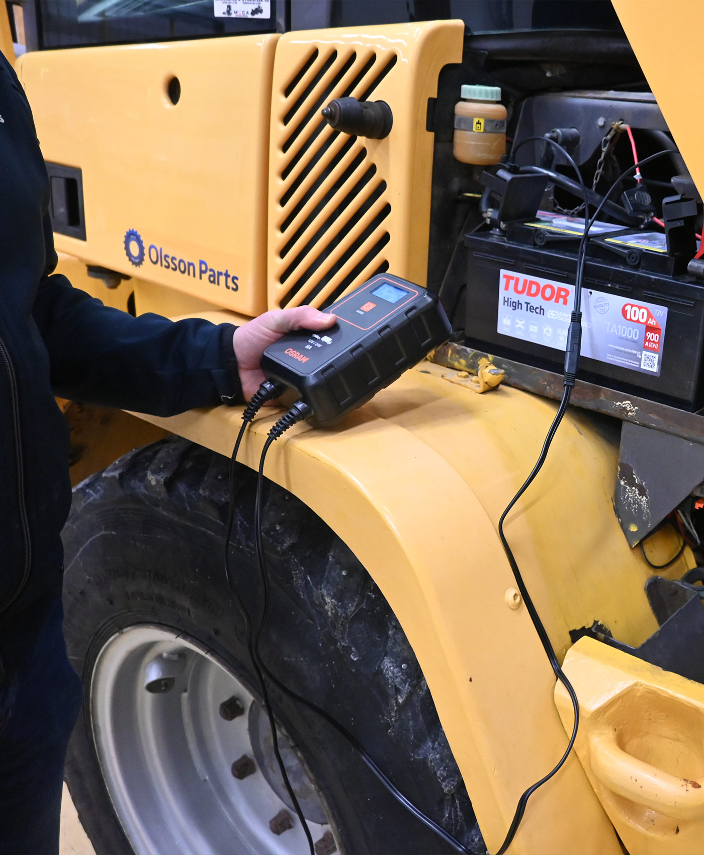 A person is holding a battery charger next to a vehicle battery on a construction machine.