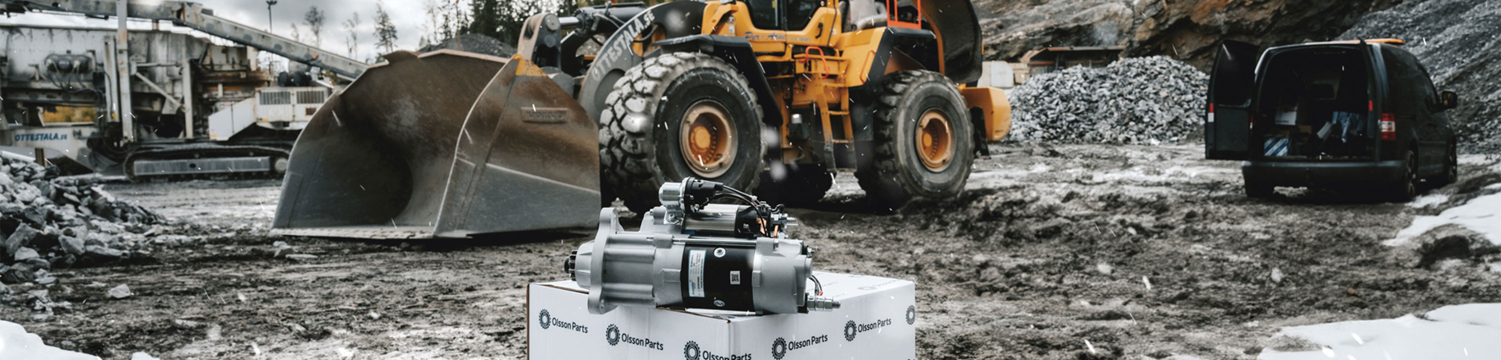 In the foreground, there is a starter motor on an Olsson Parts box. In the background, a Volvo wheel loader is standing.