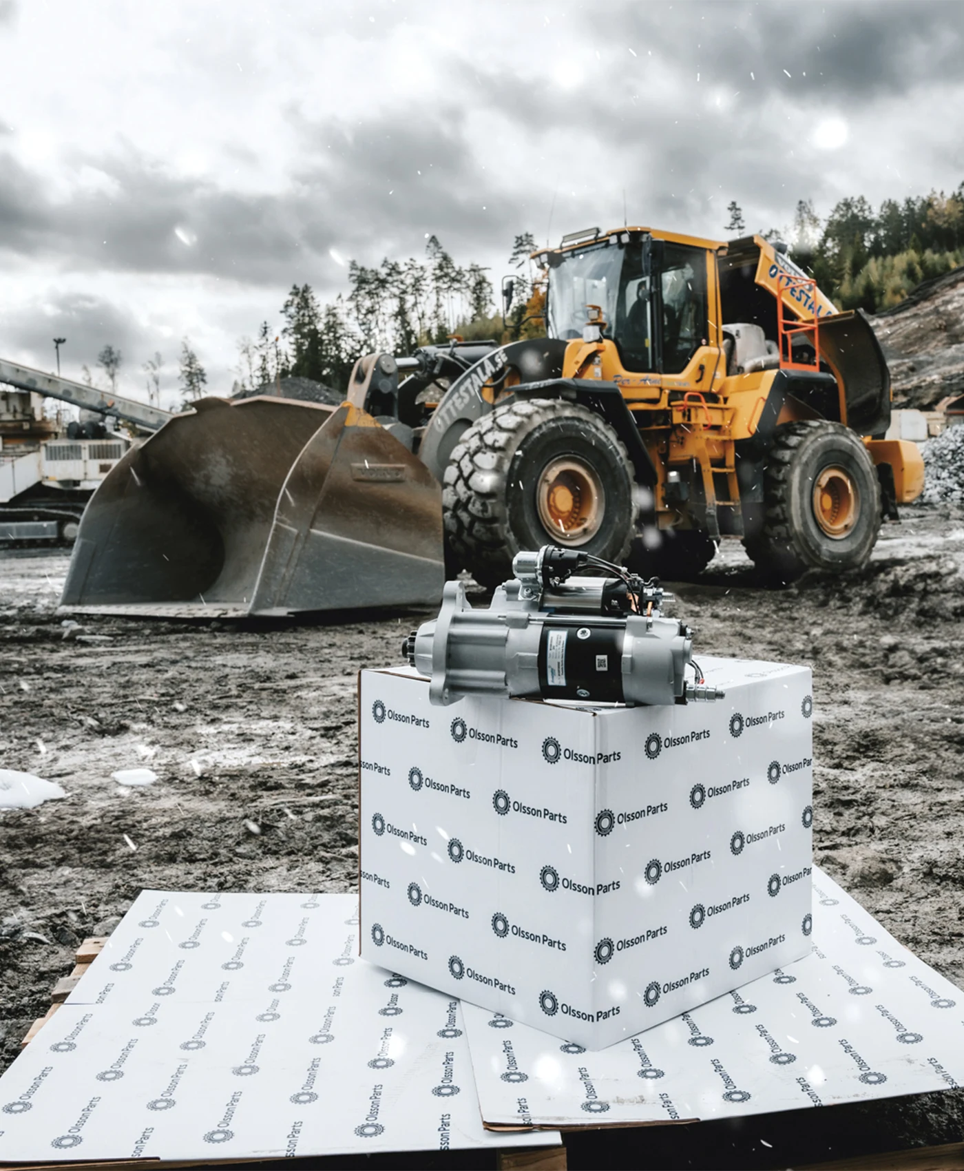 I forgrunden er en startmotor p&aring; en Olsson Parts-kasse. I baggrunden st&aring;r en Volvo-hjull&aelig;sser.