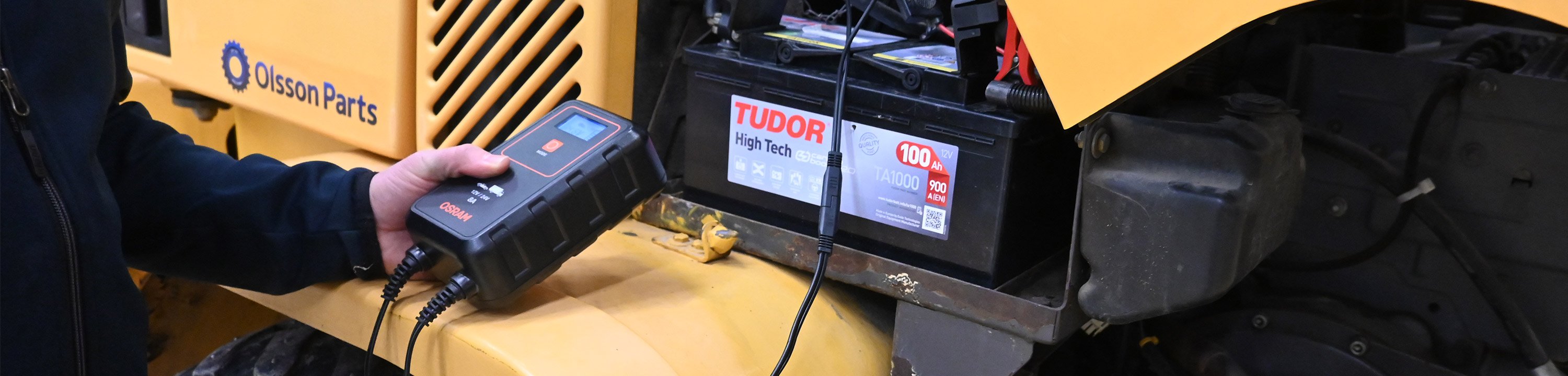 En tekniker använder en Osram-batteriladdare ansluten till ett Tudor High Tech-batteri monterat i en Volvo entreprenadmaskin.
