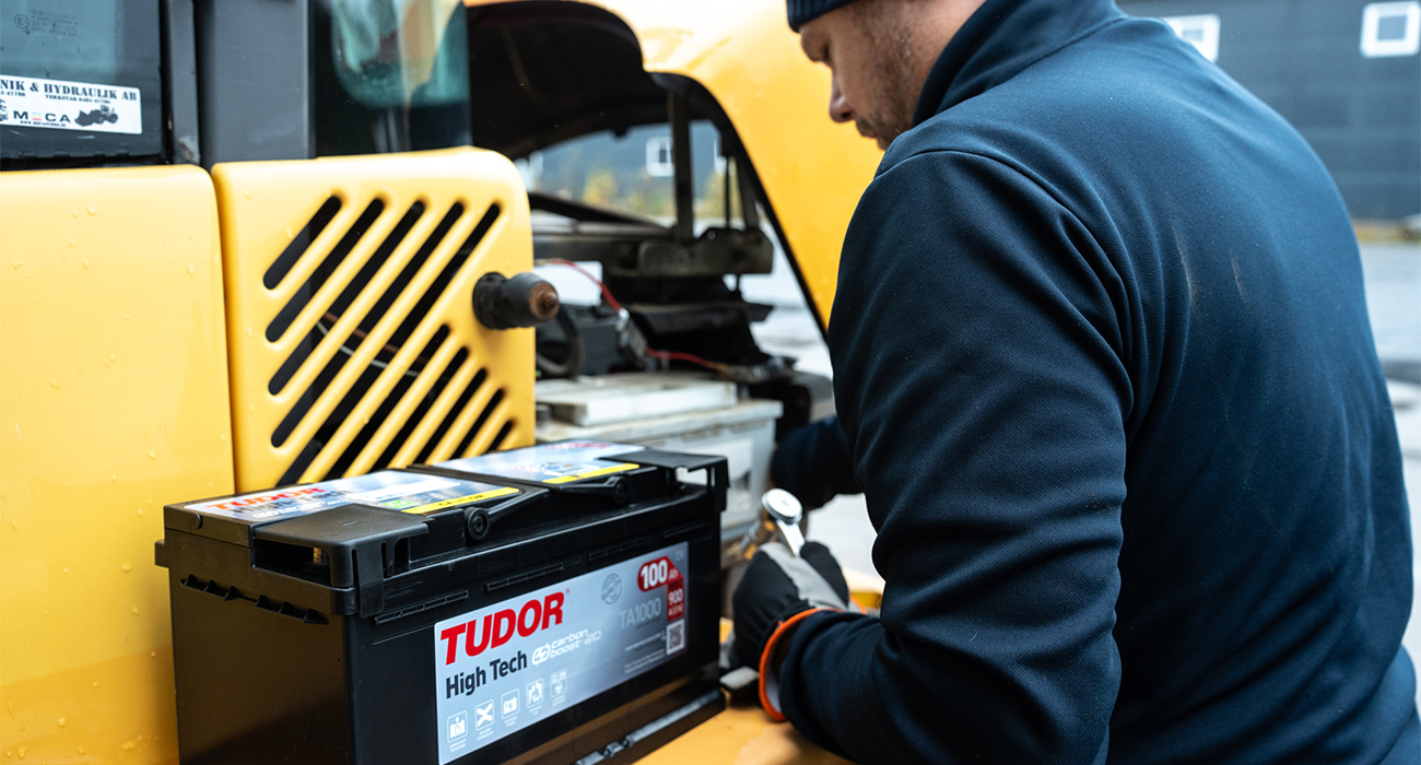 En mekaniker installerar ett Tudor High Tech-batteri på en gul entreprenadmaskin, med verktyg i handen och motorutrymmet öppet