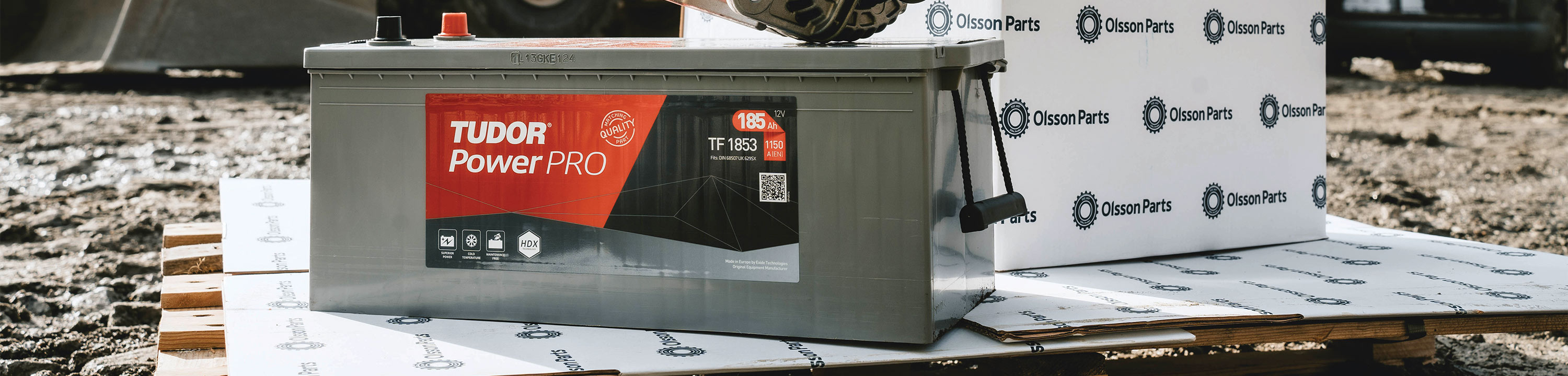 Ett stort Tudor PowerPRO-fordonsbatteri placerat på en pall utomhus, med Olsson Parts-förpackningar i bakgrunden