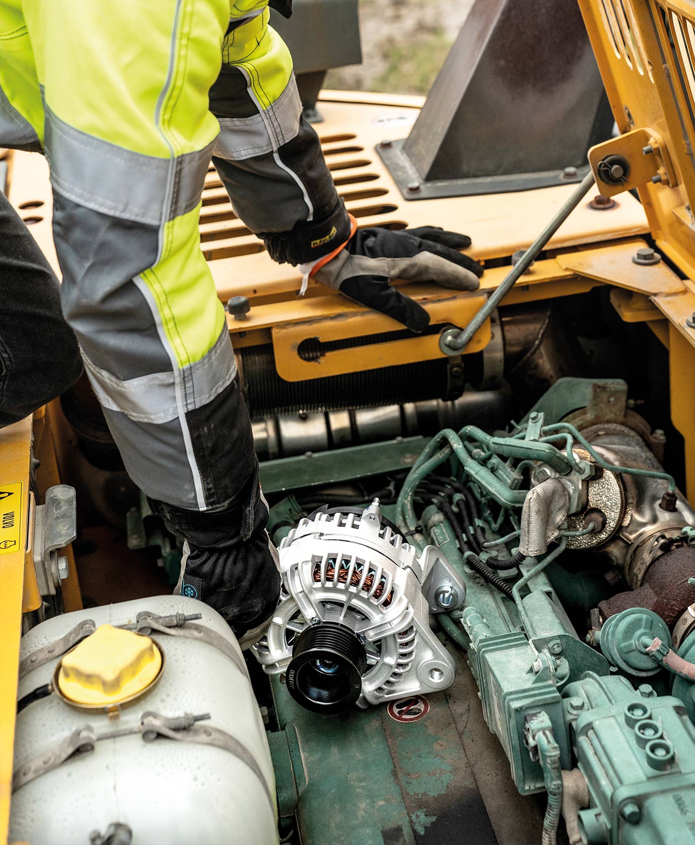 En mekaniker monterar en generator i det öppna motorutrymmet på en Volvo grävmaskin.