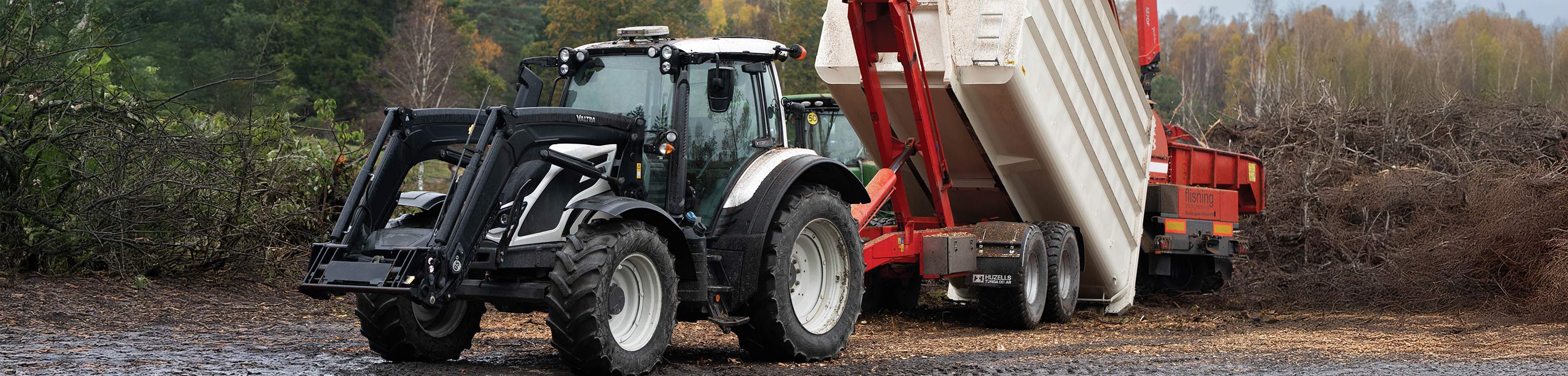En Valtra-traktor med kärra står parkerad framför en skog.