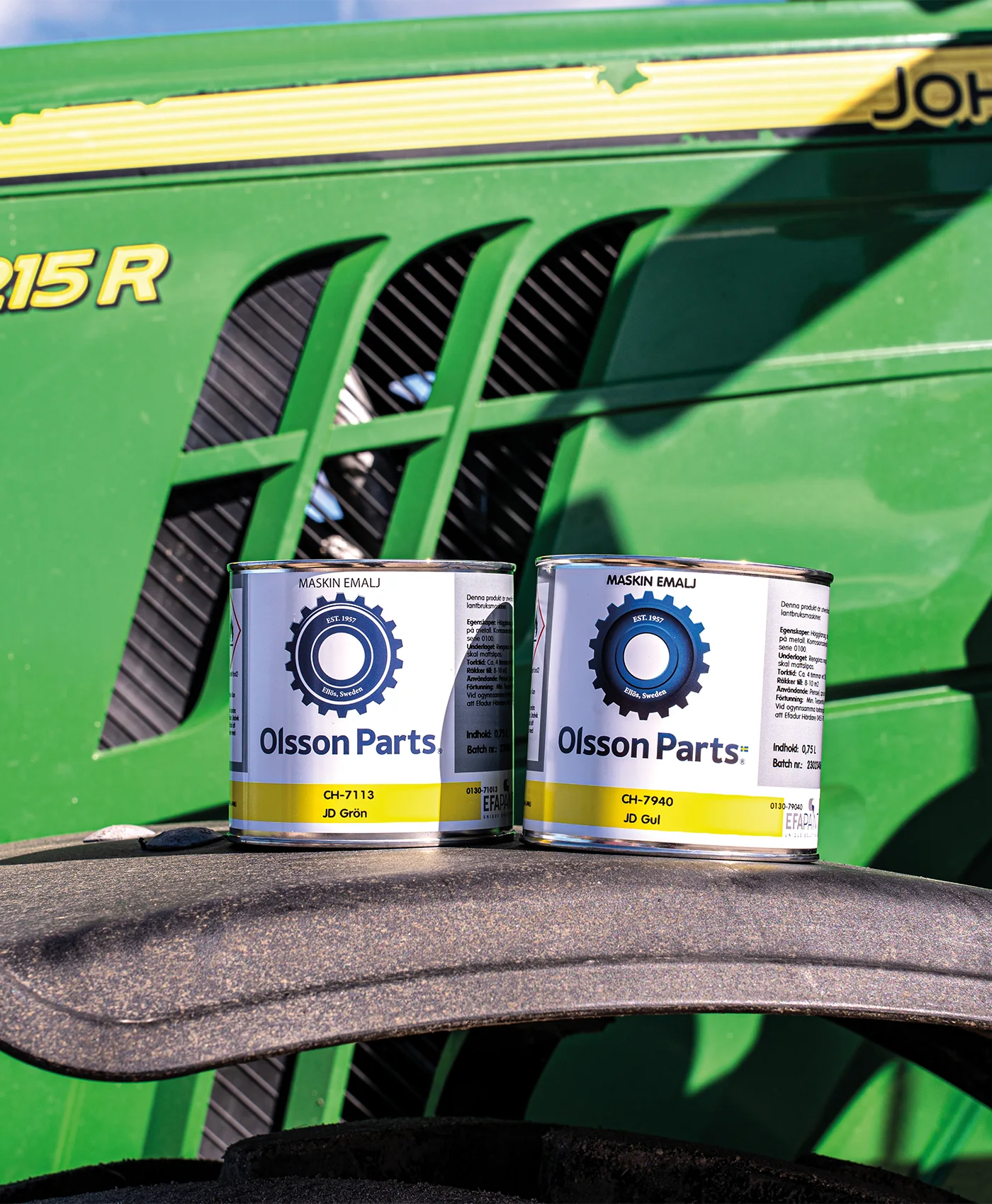 Två färgburkar från Olsson Parts står placerade på framskärmen av en John Deere-traktor.