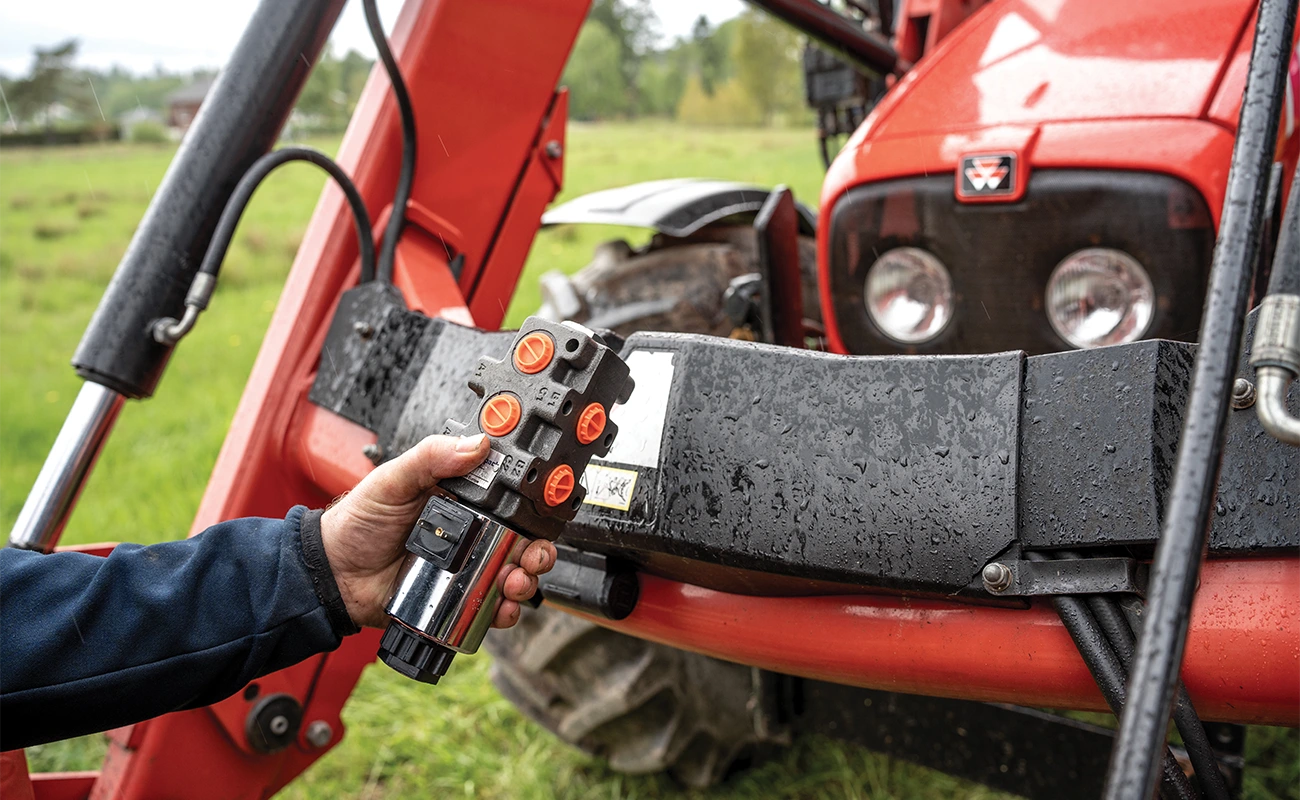 Eine Person hält ein Steuerventil vor einem Frontlader an einem Massey Ferguson Traktor.