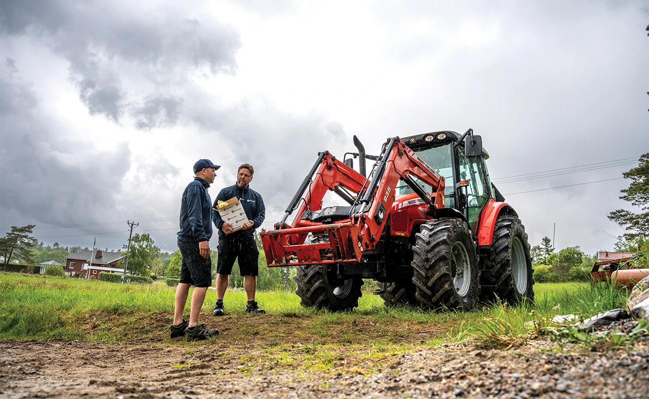To personer står foran en Massey Ferguson. Den ene holder en kasse fra Olsson Parts.