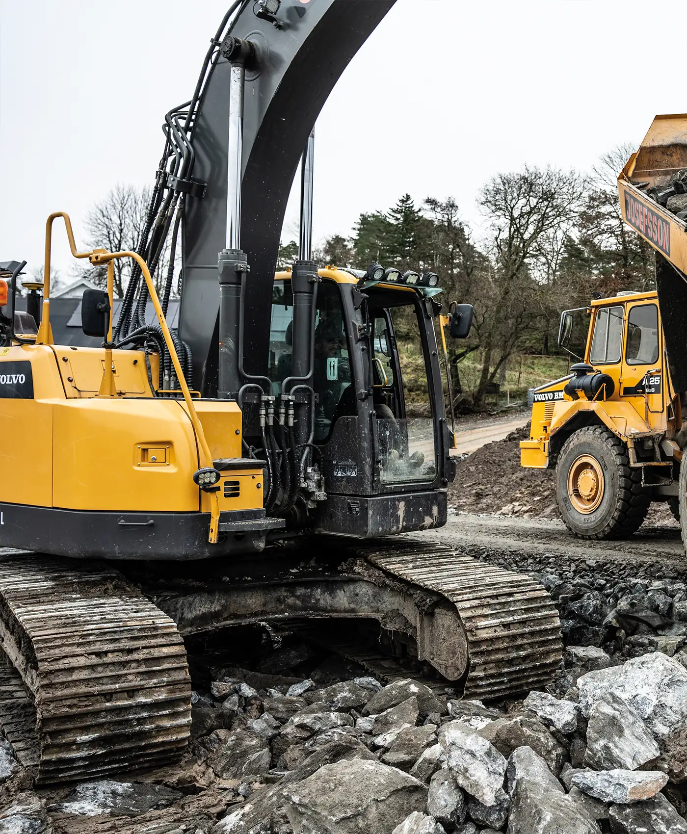 En Volvo gr&auml;vmaskin gr&auml;ver bland stenar. I bakgrunden st&aring;r en Volvo dumper parkerad.