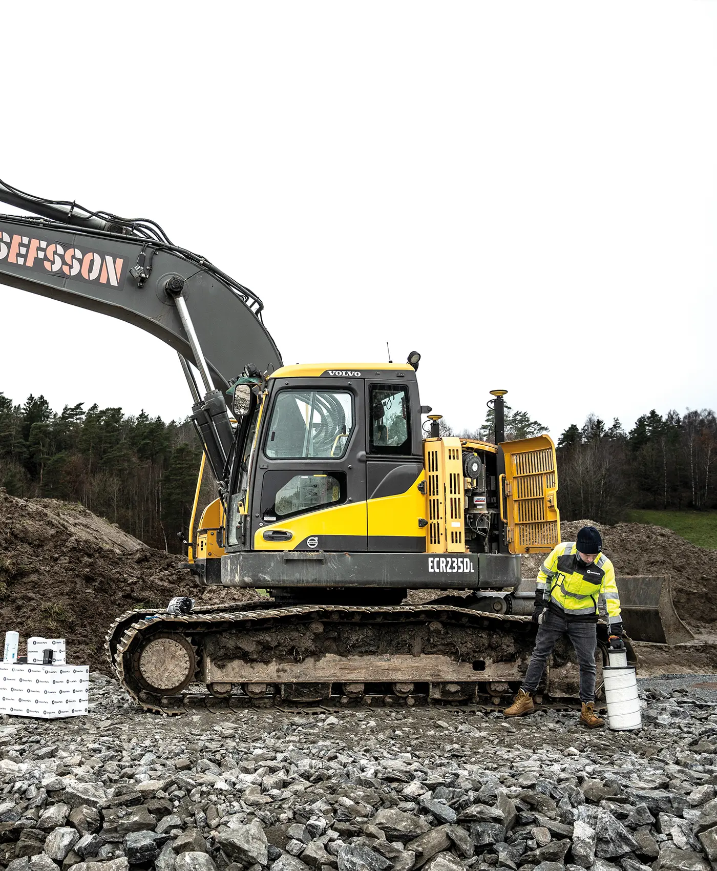 En person str&auml;cker sig efter reservdelar framf&ouml;r en Volvo gr&auml;vmaskin. Bredvid st&aring;r l&aring;dor fr&aring;n Olsson Parts.