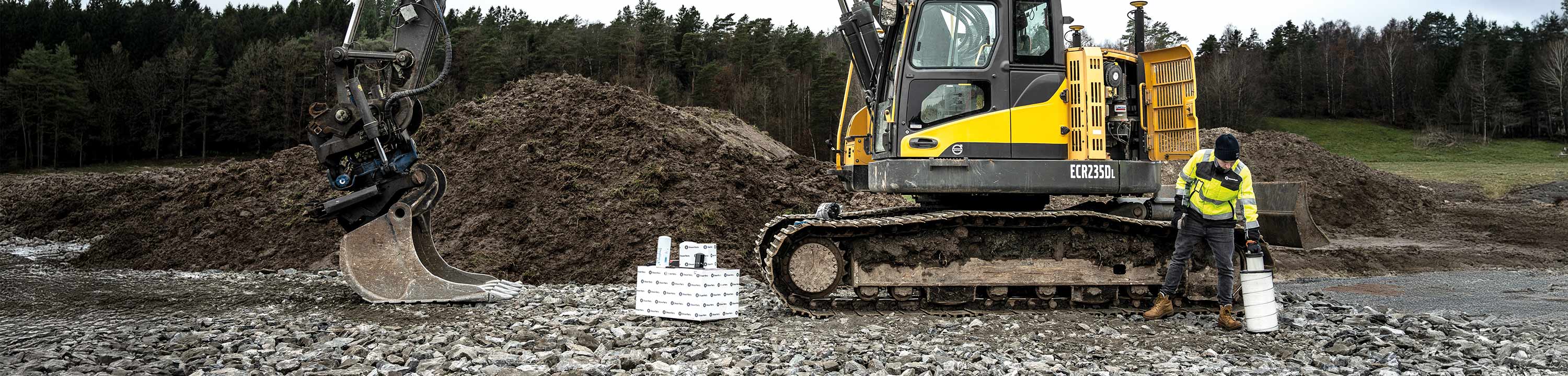 En Volvo gravemaskin med en mann gående foran. Ved siden av står en kasse med reservedeler.