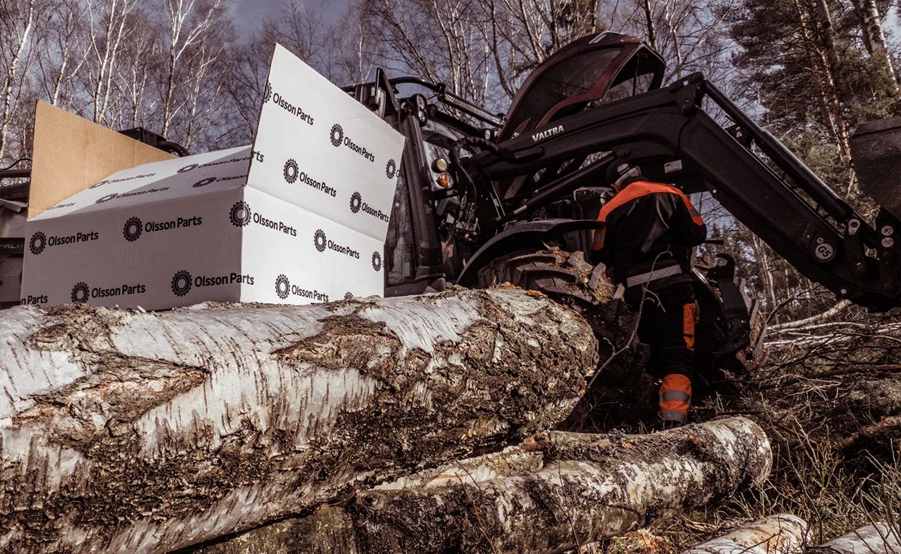 En person st&aring;r vid en Valtra traktor i skogen. I f&ouml;rgrunden st&aring;r en Olsson Parts l&aring;da placerad.